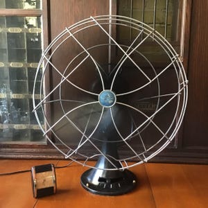 May include: An antique black electric fan with a silver wire cage and a small clock. The fan has a round base and a blue and silver emblem in the center. The clock is black and gold.