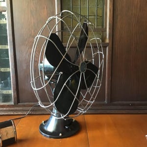 May include: A vintage black and silver electric fan with a cage-like design. The fan has four black blades and a round base. The fan is sitting on a wooden surface.