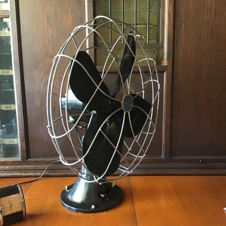 May include: A vintage black and silver electric fan with a cage-like design. The fan has four black blades and a round base. The fan is sitting on a wooden surface.