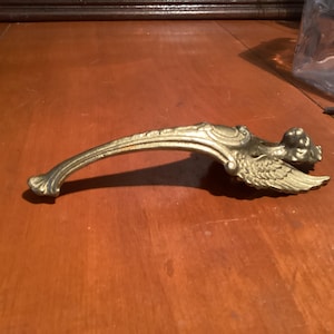 May include: A vintage brass door handle with an ornate design. The handle features a curved shape with decorative detailing, including a winged figure at one end. The brass has a warm, aged patina, suggesting its antique nature. The handle is likely for a cabinet or door.