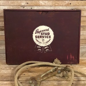 May include: A burgundy wooden box with a white and red emblem that reads "Improved Stud Service Curtis Cary Illinois Farms". A beige rope and metal clasp are in the foreground. The box has a slot for opening.