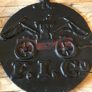 May include: Black cast iron fire mark with a red and black fire engine design. The fire engine has two large wheels and a raised platform. The text "F.I. Co." is embossed on the bottom of the mark.