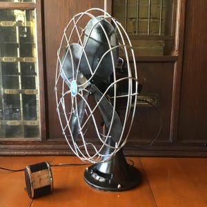 May include: An antique black electric fan with a silver wire cage and blades. The fan has a round base with brass accents and a separate motor. The fan is displayed on a wooden surface.