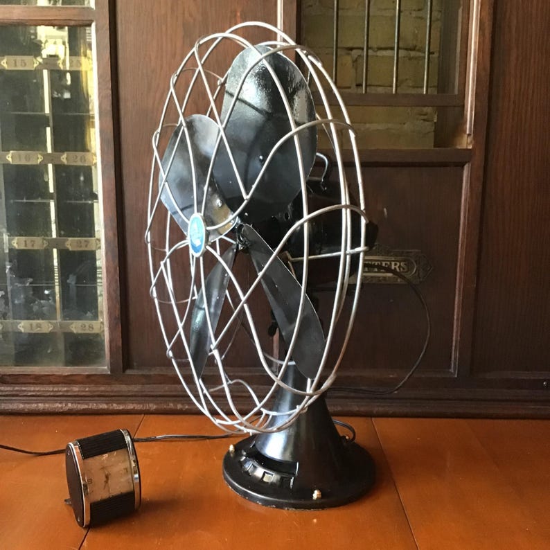 May include: An antique black electric fan with a silver wire cage and blades. The fan has a round base with brass accents and a separate motor. The fan is displayed on a wooden surface.