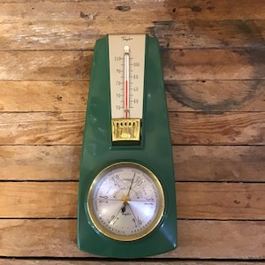 May include: A green wall-mounted Taylor weather station featuring a vertical thermometer with a gold-toned scale and a circular barometer below it. The barometer has a gold-colored rim, a white face with black text, and a central needle indicator.