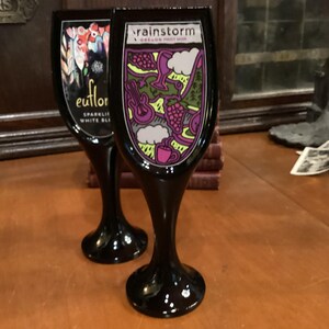 May include: Two black wine tap handles with colorful label designs. One features the text "euflor SPARKLING WHITE BLI" and floral art. The other displays "rainstorm OREGON FOOT NOR" with grapes, a guitar, and a cup.