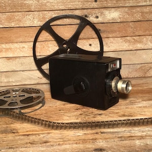 May include: A vintage black film camera with a silver lens and a black film reel. A second film reel and a strip of film are also present. The items are arranged on a wooden surface.