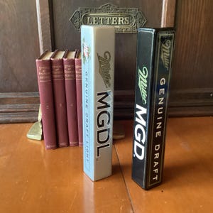 May include: Two vintage Miller MGD beer tap handles. One is light gray with blue text, the other is black with green text. The handles are rectangular and feature the Miller logo and "Genuine Draft" text. A row of red books is in the background.