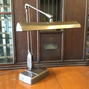 May include: A vintage desk lamp with a rectangular base and a long, adjustable arm. The lamp has a metallic finish and a rectangular shade. The lamp is positioned in front of a wooden cabinet with a "LETTERS" sign.