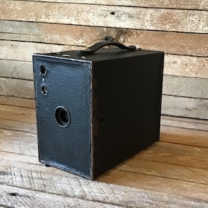 May include: A black vintage box camera with a leather covering and a carrying handle. The camera has a round lens and two small holes on the side.