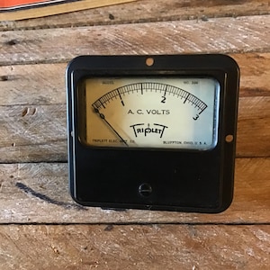 May include: A black vintage analog voltmeter with a white face and a needle pointing to 2. The face reads "A.C. VOLTS" and "TRIPLETT" with the company name "TRIPLETT ELEC. INST. CO." and "BLUFFTON, OHIO U.S.A."