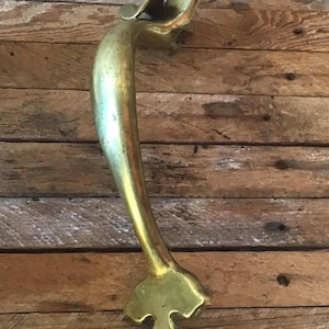 May include: A vintage brass door handle with a decorative, ornate design. The handle is in the shape of a curved arch with a pointed tip at the bottom.