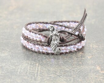 May include: A wrap bracelet featuring light purple beads and brown leather straps. A silver mermaid charm is centered on the bracelet. The bracelet is designed to wrap multiple times around the wrist.