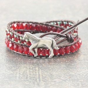 May include: A red and brown leather wrap bracelet with a silver horse charm. The bracelet is made with red glass beads and has a leather tie closure.
