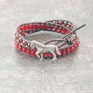 May include: A red and brown leather wrap bracelet with a silver horse charm. The bracelet is made with small red glass beads and has a leather tie closure.
