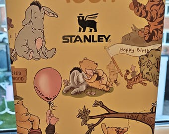 May include: A rectangular cardboard box with a light yellow background featuring Winnie the Pooh characters. The Stanley logo is centred with the text Winnie the Pooh above it. Illustrations include Pooh, Piglet, Tigger, Eeyore, and Christopher Robin with signs reading Bounce This Way, Happy Birthday, and 100 Aker Wood.