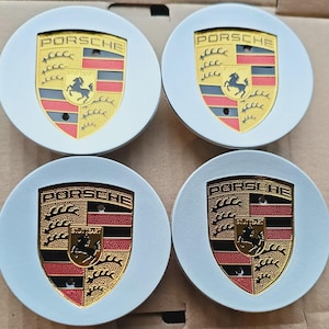 May include: Four circular silver wheel centre caps, each featuring a centred gold, red, and black Porsche crest emblem. The crest includes the word PORSCHE at the top, a black horse in the centre, and the word STUTTGART on a small gold shield.