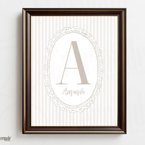 May include: A framed print with a dark brown frame and gold inner edge. The print features a light taupe letter "A" inside a decorative oval frame, with the name "Amanda" below. The background has vertical stripes.