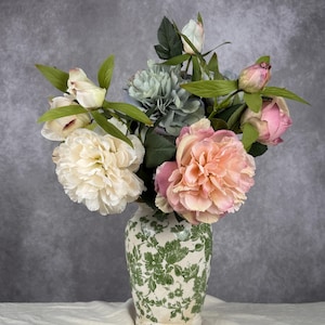 May include: A floral display in a patterned vase. The bouquet features cream peonies, peach-toned flowers, and green leaves. The vase, with a green floral design, sits on a white cloth against a grey background.