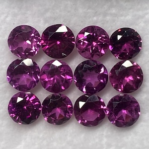 Puede incluir: Una colección de doce piedras preciosas redondas, facetadas y de color magenta intenso, dispuestas en una cuadrícula de tres por cuatro dentro de una caja de presentación blanca. Cada piedra presenta un corte brillante con múltiples facetas.