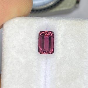 May include: A rectangular, faceted pink gemstone displayed on a white foam insert. A metal dial caliper is positioned in the background, showing a measurement of approximately 0.25 inches on the gauge face for the stone's dimensions.