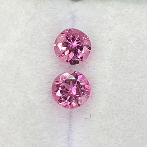 Puede incluir: Dos gemas rosas de corte redondo y brillante se muestran sobre una superficie blanca. Las gemas tienen un color vibrante y saturado y probablemente sean diamantes u otras piedras preciosas. Las gemas están sostenidas por los dedos de una persona.