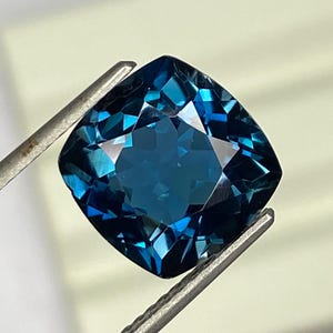 Puede incluir: Una gema de color verde azulado oscuro, tallada en forma de cojín, es sostenida por unas pinzas plateadas sobre un fondo blanco suave. Las facetas de la gema reflejan la luz, mostrando su talla brillante.
