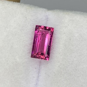 May include: A rectangular, faceted, hot pink gemstone. The gem is held on a white surface. The gem has a bright, reflective surface and is likely a cut stone for jewelry.