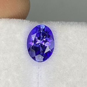 May include: An oval-shaped, deep blue gemstone. The gem is displayed on a white, textured surface. The stone's facets reflect light, creating a sparkling effect. The gemstone is a vibrant shade of blue.