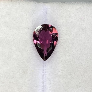 Peut inclure: Une pierre précieuse taillée en forme de poire, de couleur magenta foncé. La gemme est posée sur un fond blanc, avec un instrument de mesure partiellement visible. La forme et la couleur suggèrent une grenat ou une tourmaline.