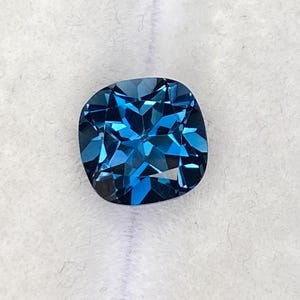 Op de afbeelding: Een kussengeslepen, diepblauwe edelsteen wordt getoond op een witte ondergrond. De edelsteen heeft een gefacetteerd oppervlak dat licht reflecteert, waardoor een sprankelend effect ontstaat. Een streng blauwe kralen is zichtbaar op de achtergrond.