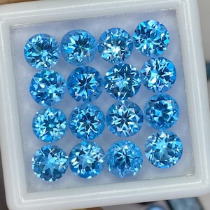 Op de afbeelding: Een verzameling van zestien ronde, gefacetteerde, lichtblauwe edelstenen, gerangschikt in een witte vierkante container. Extra losse edelstenen en gereedschap zijn zichtbaar op de achtergrond. De edelstenen hebben een briljant geslepen.