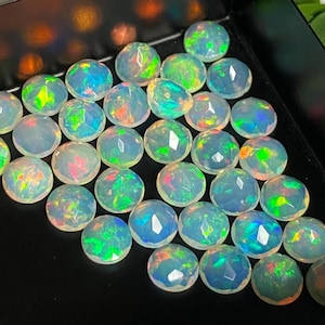 May include: A selection of round, faceted opals in a black tray. The opals exhibit a spectrum of colours, including blue, green, and red, with a shimmering, iridescent effect. The gemstones are closely arranged.