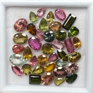 Könnte beinhalten: Eine Sammlung farbenfroher Edelsteine in verschiedenen Formen, darunter oval, tropfenförmig und rechteckig. Die Edelsteine werden in einer weißen Schale präsentiert und zeigen eine Farbpalette von Rosa und Grün bis hin zu Gelb und Braun.