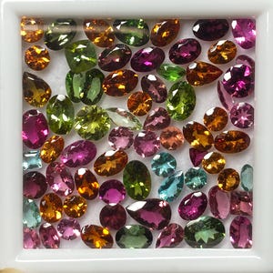 May include: A collection of colorful gemstones in a white square container. The gems are various shapes and sizes, including oval, pear, and round cuts. Colors range from pink, green, orange, and blue. The gemstones are displayed against a white background.