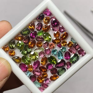 May include: A collection of colorful gemstones in various shapes and sizes, including oval, pear, and round cuts. The gems are displayed in a white square tray, showcasing a range of colors from green and blue to pink and orange.