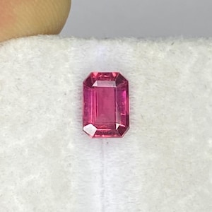 Puede incluir: Una gema de color rosa vibrante con corte esmeralda sobre una almohadilla de terciopelo blanco. Un calibrador mecánico está colocado sobre la piedra, mostrando una medida de aproximadamente 0,6 cm en el dial para la gema suelta.