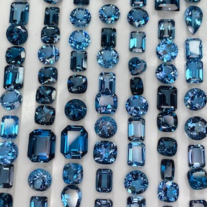 Op de afbeelding: Een verzameling blauwe edelstenen in verschillende vormen, waaronder rond, ovaal en vierkant, gepresenteerd op een witte ondergrond. De edelstenen hebben verschillende blauwtinten en zijn waarschijnlijk bedoeld voor het maken van sieraden.