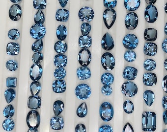 May include: An assortment of blue gemstones in various shapes, including oval, pear, and square cuts, arranged in vertical rows. The gems have a translucent quality and a range of blue hues, from light to deep shades.