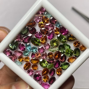 May include: A collection of colorful gemstones in a white square display case. The gemstones are various shapes and sizes, including oval, pear, and round cuts. Colors include pink, green, orange, and blue. The gemstones are arranged on a white surface.