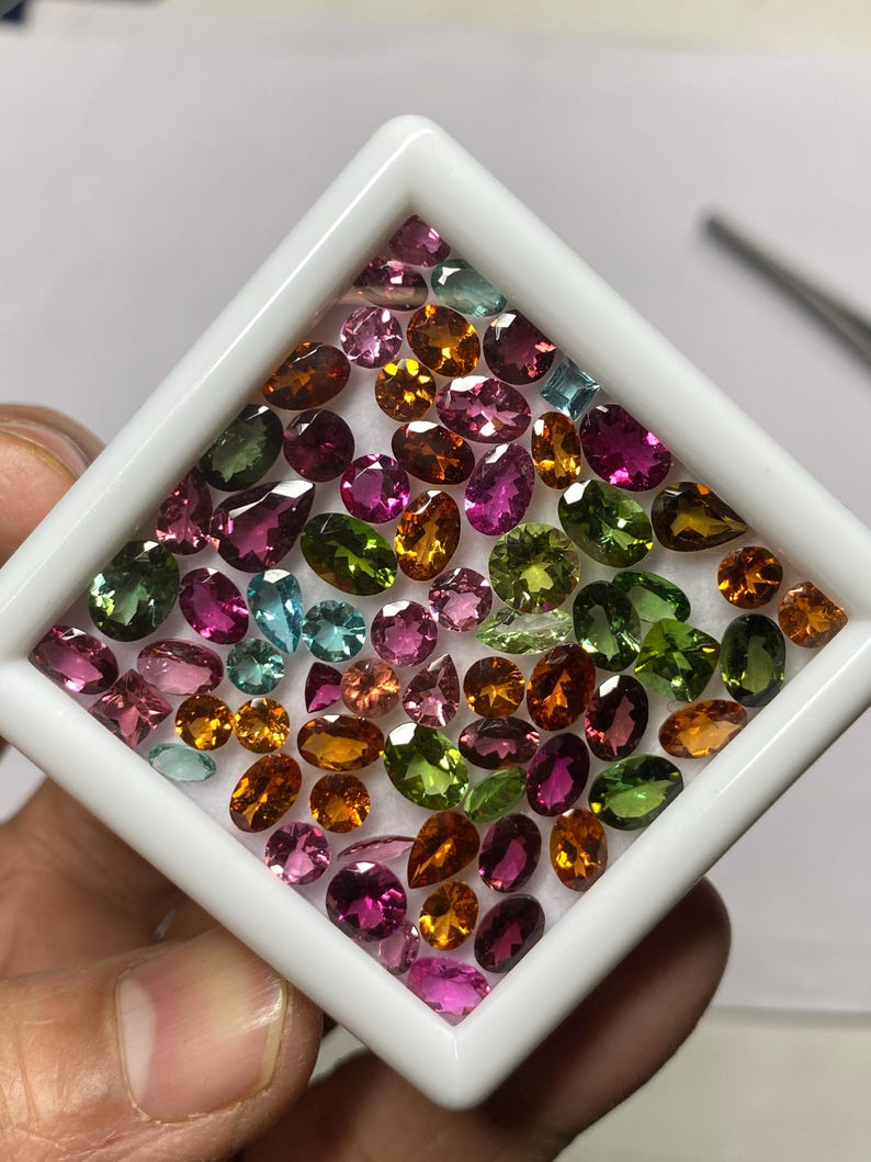 May include: A collection of colorful gemstones in a white square display case. The gemstones are various shapes and sizes, including oval, pear, and round cuts. Colors include pink, green, orange, and blue. The gemstones are arranged on a white surface.