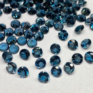 May include: A selection of round, faceted gemstones in a deep teal hue. The gems, of varying sizes, are scattered across a white surface. The facets of each stone reflect light, creating a sparkling effect. The gemstones are likely for jewellery making.