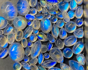Puede incluir: Una colección de piedras preciosas de luna pulidas en diversas formas, incluyendo óvalos y lágrimas. Las piedras exhiben una cautivadora adularescencia azul, con destellos de color. Las gemas están dispuestas sobre una superficie oscura.