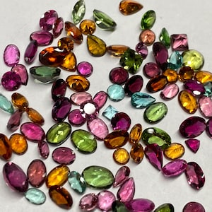 May include: A collection of loose gemstones in various shapes and colors. The gems include oval, pear, and square cuts in shades of pink, green, orange, yellow, and blue. The gemstones are scattered across a neutral surface.