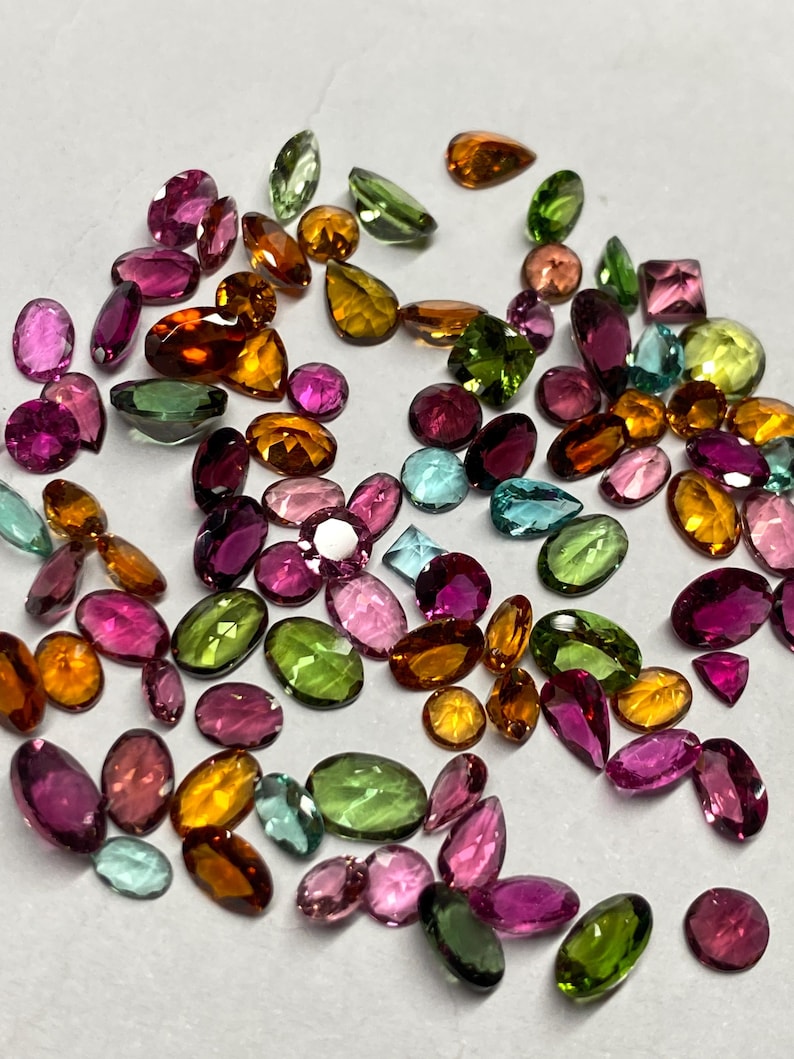 May include: A collection of loose gemstones in various shapes and colors. The gems include oval, pear, and square cuts in shades of pink, green, orange, yellow, and blue. The gemstones are scattered across a neutral surface.