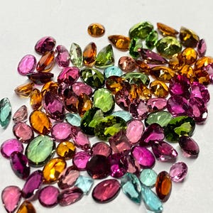May include: A collection of faceted gemstones in various shapes and sizes. The gems display a range of colors, including pink, green, orange, and blue. The gemstones are scattered on a white surface.