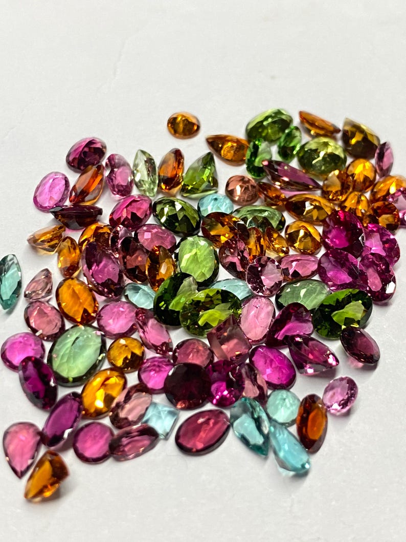 May include: A collection of faceted gemstones in various shapes and sizes. The gems display a range of colors, including pink, green, orange, and blue. The gemstones are scattered on a white surface.
