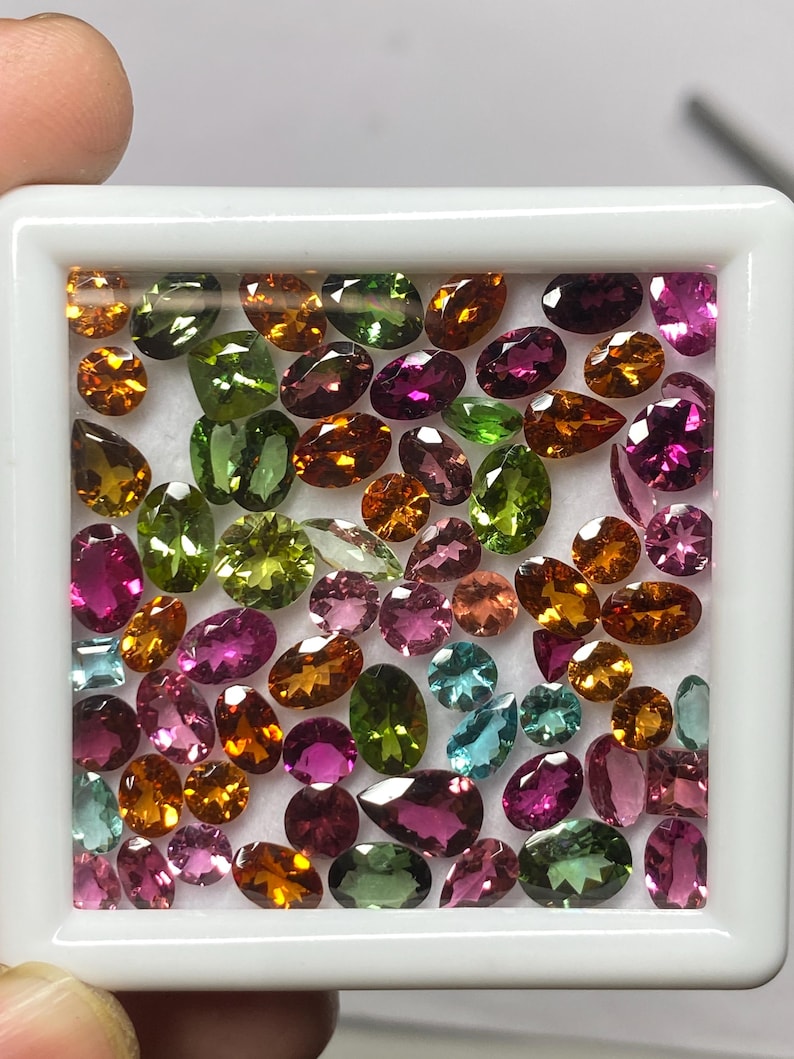 May include: A collection of colorful gemstones in a white square container. The gems are various shapes and sizes, including oval, pear, and round cuts. Colors range from pink, green, orange, and blue. The gemstones are displayed against a white background.