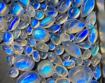 May include: A collection of polished moonstone gemstones in various shapes and sizes. The stones display a captivating blue and iridescent sheen, with a translucent appearance. The gemstones are arranged on a dark surface, showcasing their natural beauty.
