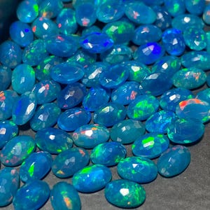 May include: A collection of faceted, oval-shaped opals. The opals are a vibrant blue color, with flashes of green, red, and orange. The gemstones are displayed closely together, filling the frame.